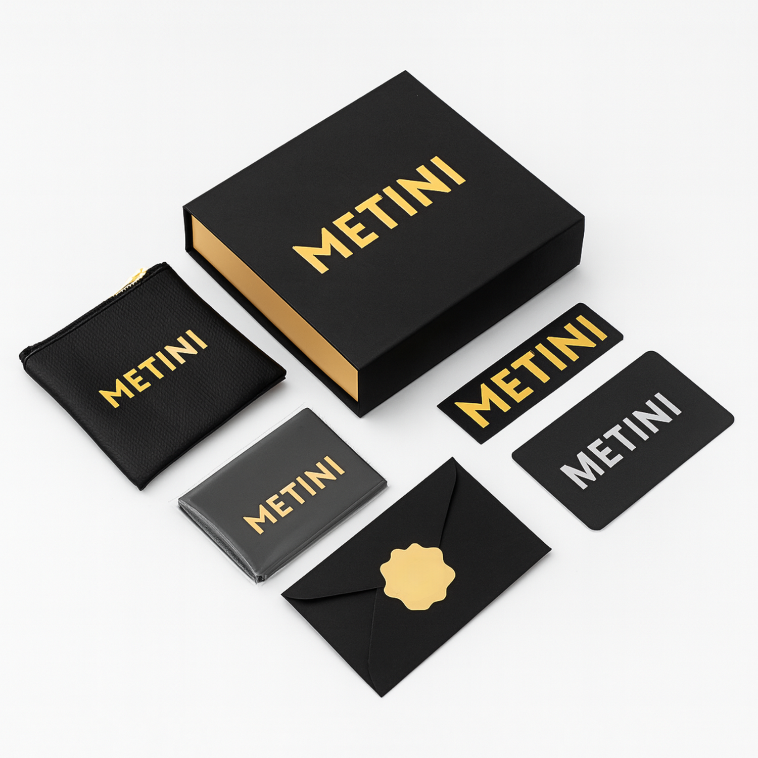 Opakowanie prezentowe METINI z czarnym pudełkiem, etui i elementami z logo w złotym kolorze: naklejka, koperta z kartą gwarancyjną, ściereczka z logo z mikrofibry i saszetka na suwak z ekoskóry.